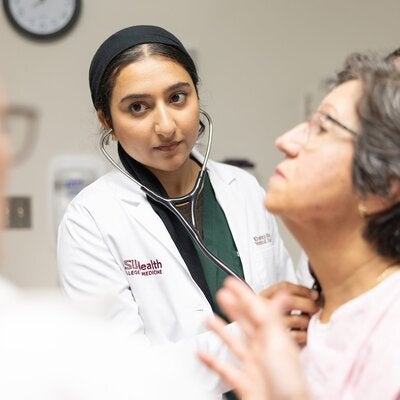 A doctor in an FSUHealth lab coat uses a stethoscope on a female patient.